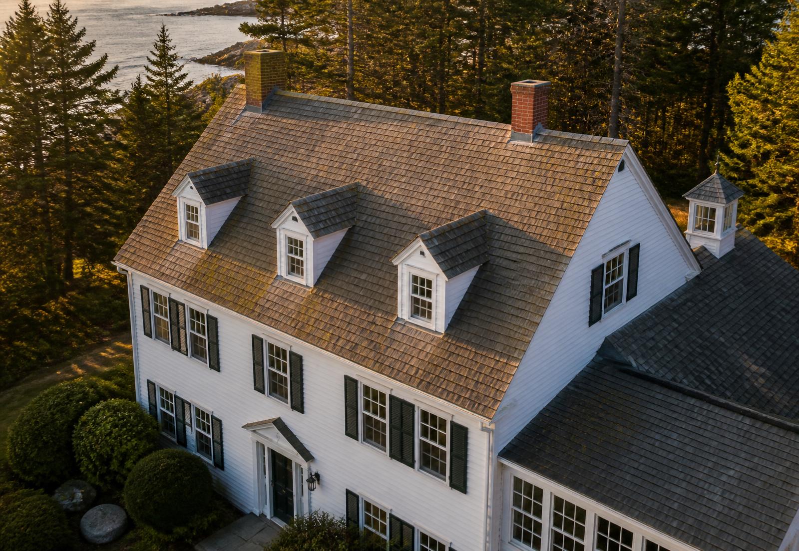 Aerial view of a coastal Maine colonial home with an aging asphalt shingle roof at golden hour